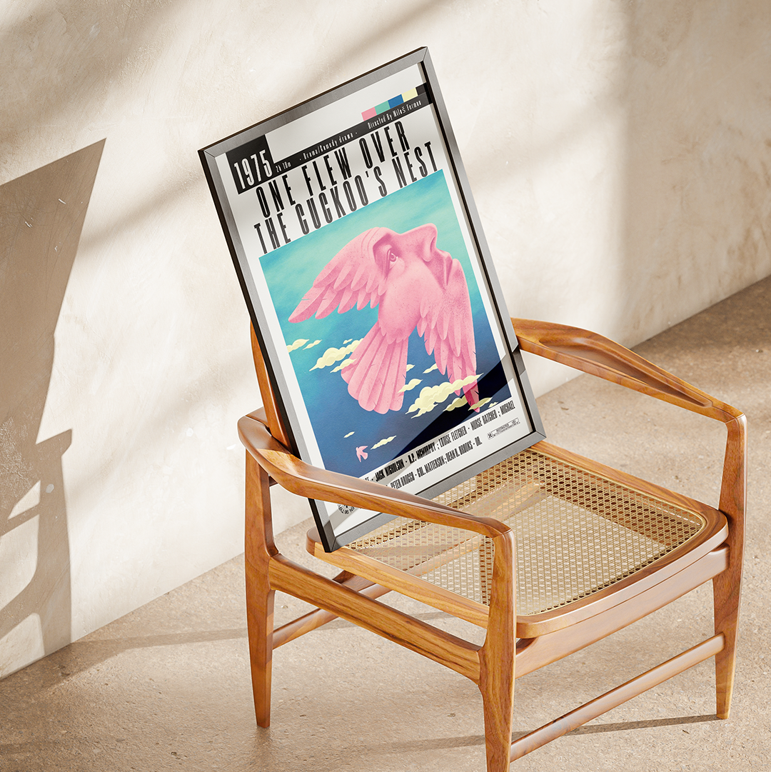 A framed art print featuring a pink bird and the text '1975 - WE'VE GOT TO GET TO THE TOP OF THE HILL' is propped against a wooden cane-seated chair in a sunlit room with a textured beige wall.