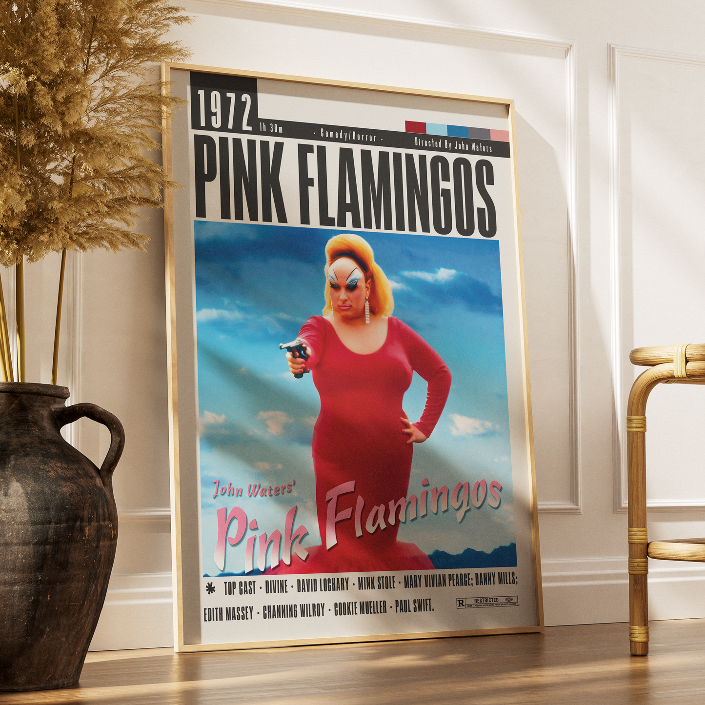 A framed movie poster for 'Pink Flamingos' leaning against a white wall on a wooden floor, styled in a room with a dark ceramic vase and dried grass on the left and a gold chair on the right.