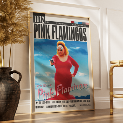 A framed movie poster for 'Pink Flamingos' leaning against a white wall on a wooden floor, styled in a room with a dark ceramic vase and dried grass on the left and a gold chair on the right.