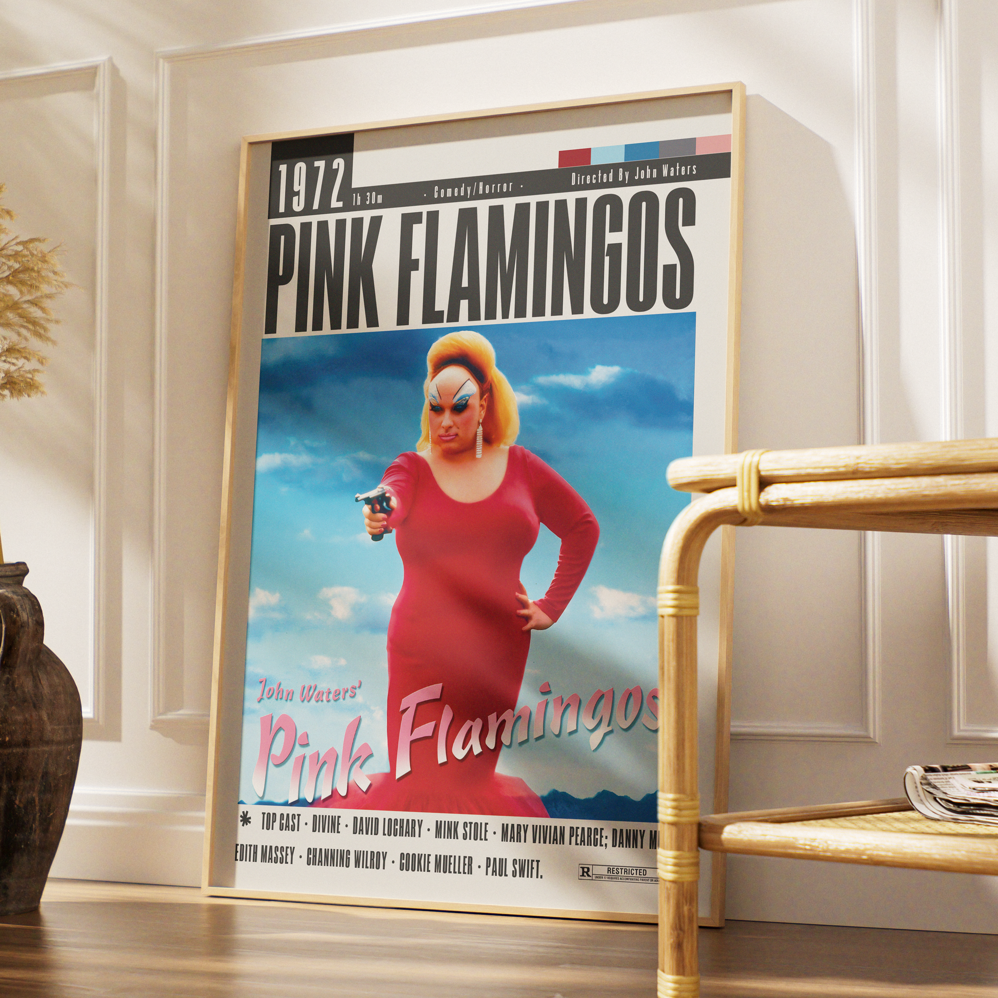 A framed vintage-style poster leaning against a white paneled wall on a wooden floor, next to a bamboo side table. The poster reads '1972 PINK FLAMINGOS' and features a woman in a red dress against a blue sky background.