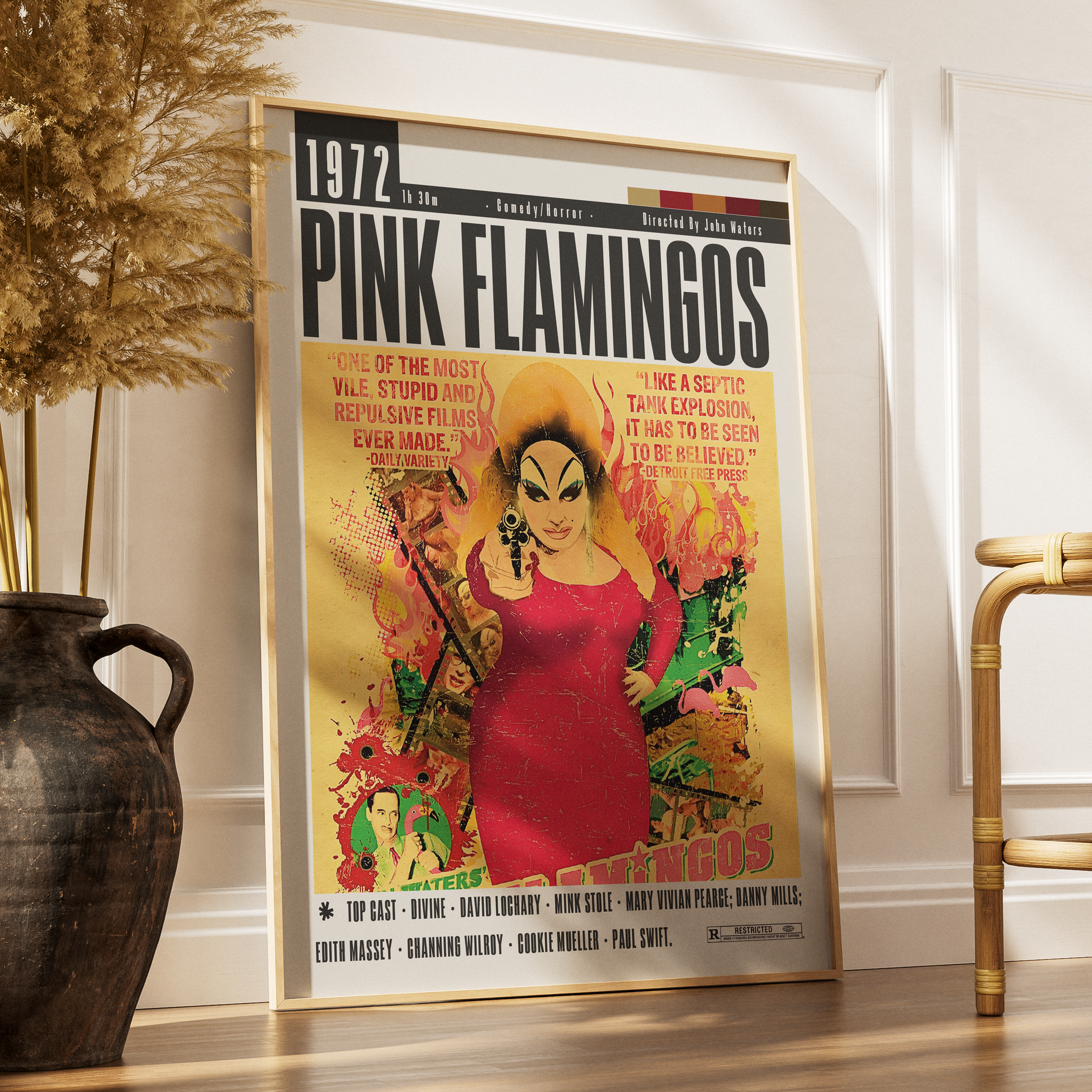 A framed vintage-style poster for Pink Flamingos leaning against a white wall on a wooden floor, with a dark vase and part of a chair visible in the room.