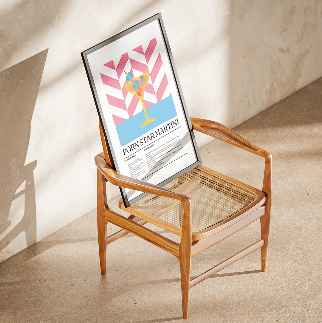 A framed poster with a colorful cocktail illustration and the text 'PORN STAR MARTINI' leans against a mid-century modern wooden armchair in a sunlit room with a textured beige wall and floor.