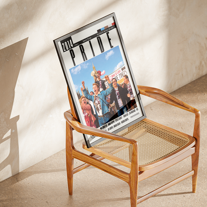 A framed 2017 PRIDE magazine leaning on a mid-century modern wooden armchair with a cane seat, set against a textured beige plaster wall with soft sunlight and shadows.