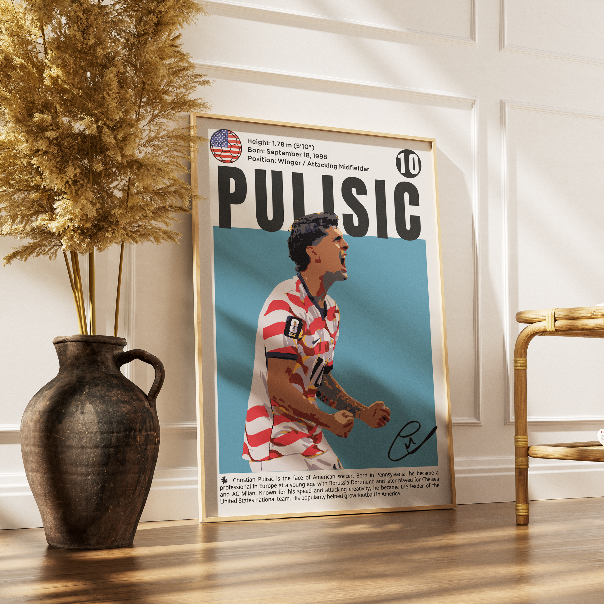 A framed poster featuring a stylized portrait of footballer Alexis Sanchez under the name 'PULISIC' with a light blue background, leaning against a white paneled wall on a wooden floor next to a dark ceramic vase with dried grass and a gold chair.