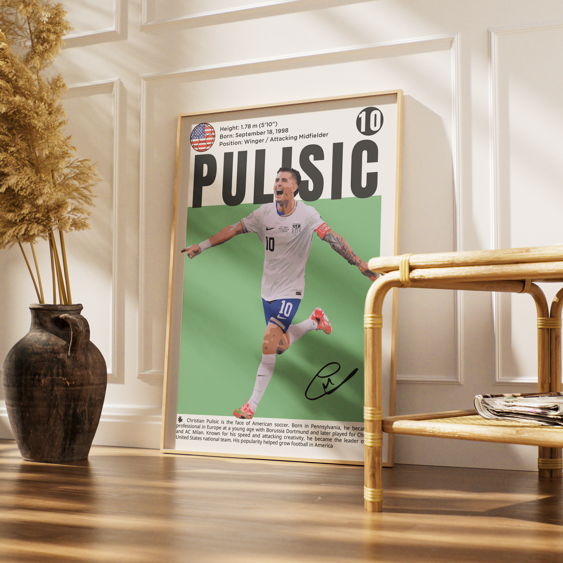 A framed poster of soccer player Curtis Pulisic leaning against a white paneled wall on a wooden floor, next to a rattan side table and a dark ceramic vase with dried grass.