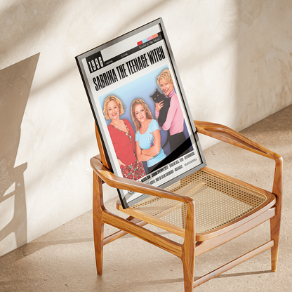 A framed magazine or book cover leaning against a wooden cane-seated chair in a sunlit room. The cover reads '1990' and 'SABRINA THE TEENAGE WITCH' with a group photo on the front.