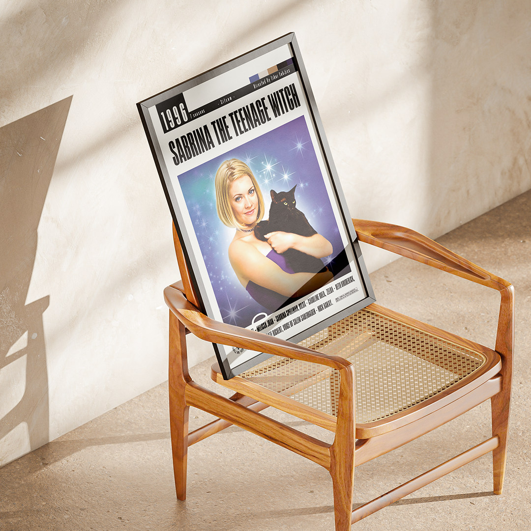 A framed art print featuring a magazine cover story titled 'SABRINA THE TEENAGE WITCH' is propped up in a mid-century modern wooden armchair with a cane seat. The scene is set in a warm, sunlit room with textured plaster walls and floor, casting soft shadows.