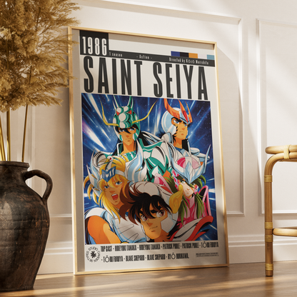 A framed Saint Seiya 1986 anime poster leaning against a white paneled wall on a wooden floor, with a dark vase and dried grass on the left and a bamboo chair on the right.