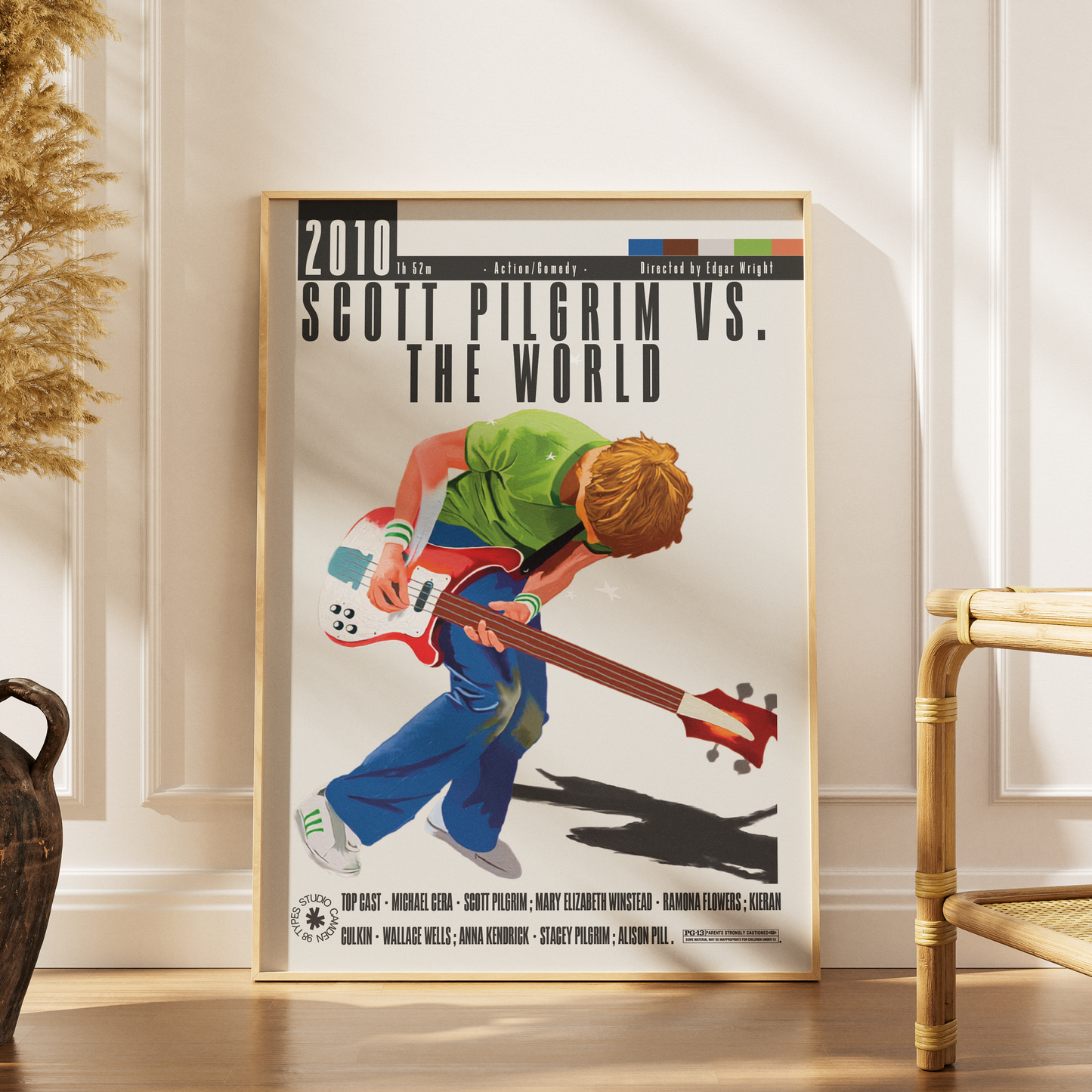 A framed poster print leaning against a white paneled wall on a wooden floor, showing an illustrated scene of Scott Pilgrim playing an electric bass guitar with the text '2010 Scott Pilgrim vs. The World.
