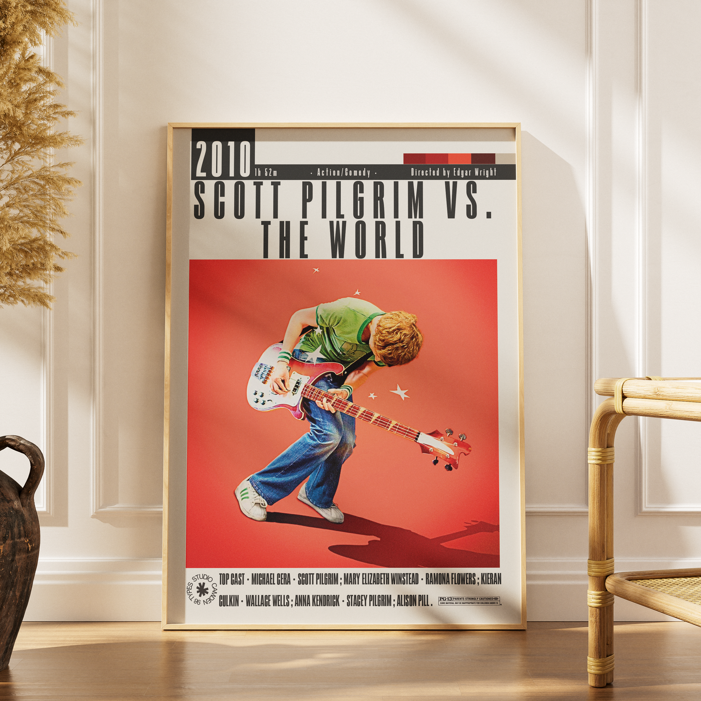 A framed poster print leaning against a white paneled wall on a wooden floor, showing a stylized illustration of Scott Pilgrim playing an electric guitar against a red background. The poster includes the text '2010 SCOTT PILGRIM VS.