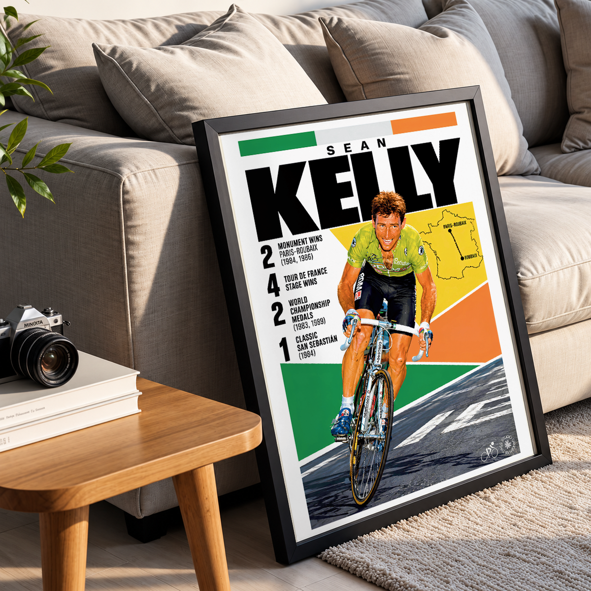 A framed cycling poster leaning against a beige sofa in a living room setting. The poster features a vintage-style illustration of Sean Kelly on a bicycle with bold text reading 'SEAN KELLY' and race statistics.
