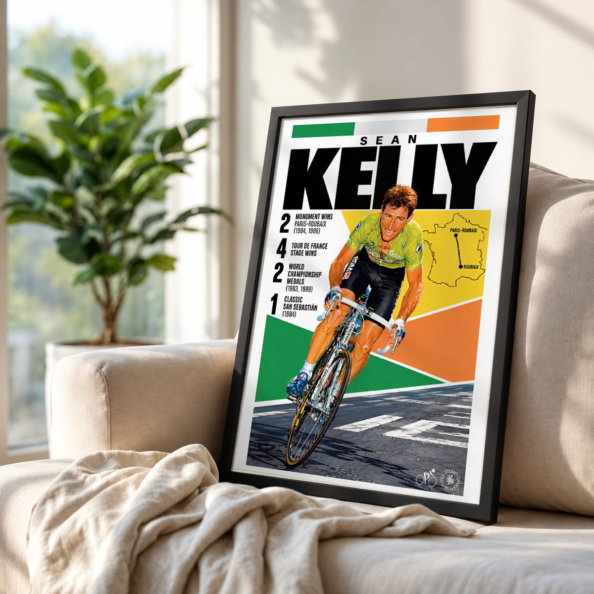 A framed cycling poster featuring Sean Kelly in a green and black cycling jersey, leaning against a beige sofa in a bright living room with a plant in the background.