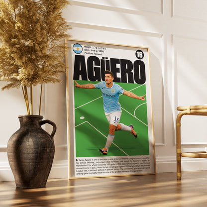A framed vertical poster of a vintage football magazine cover featuring Agüero in a light blue Manchester City kit, leaning against a white paneled wall on a wooden floor next to a dark ceramic vase with dried grass and a gold chair.