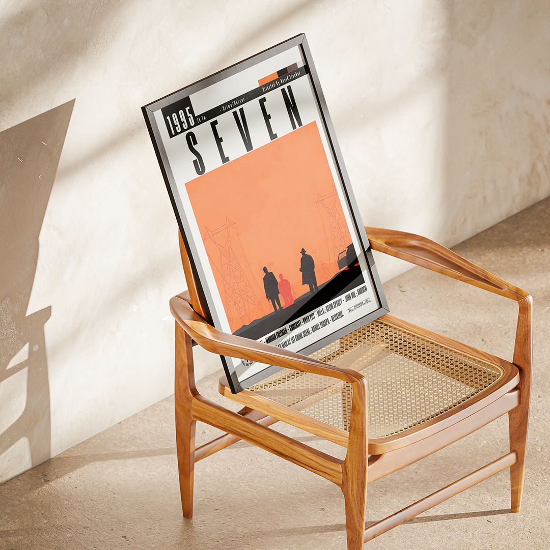 A framed art print featuring the movie poster for 'Seven' (1999) is propped up on a mid-century modern wooden armchair with a woven cane seat. The scene is styled in a warm, minimalist room with textured plaster walls and floor, lit by natural sunlight casting soft shadows.