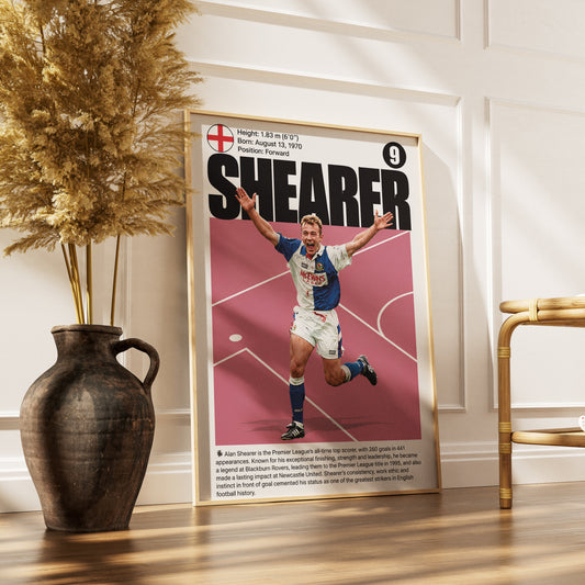 A framed vintage-style football poster leaning against a white paneled wall on a wooden floor, next to a dark vase with dried grass and a gold chair. The poster features the name SHEARER in large black letters over a pink field with a football player in action.