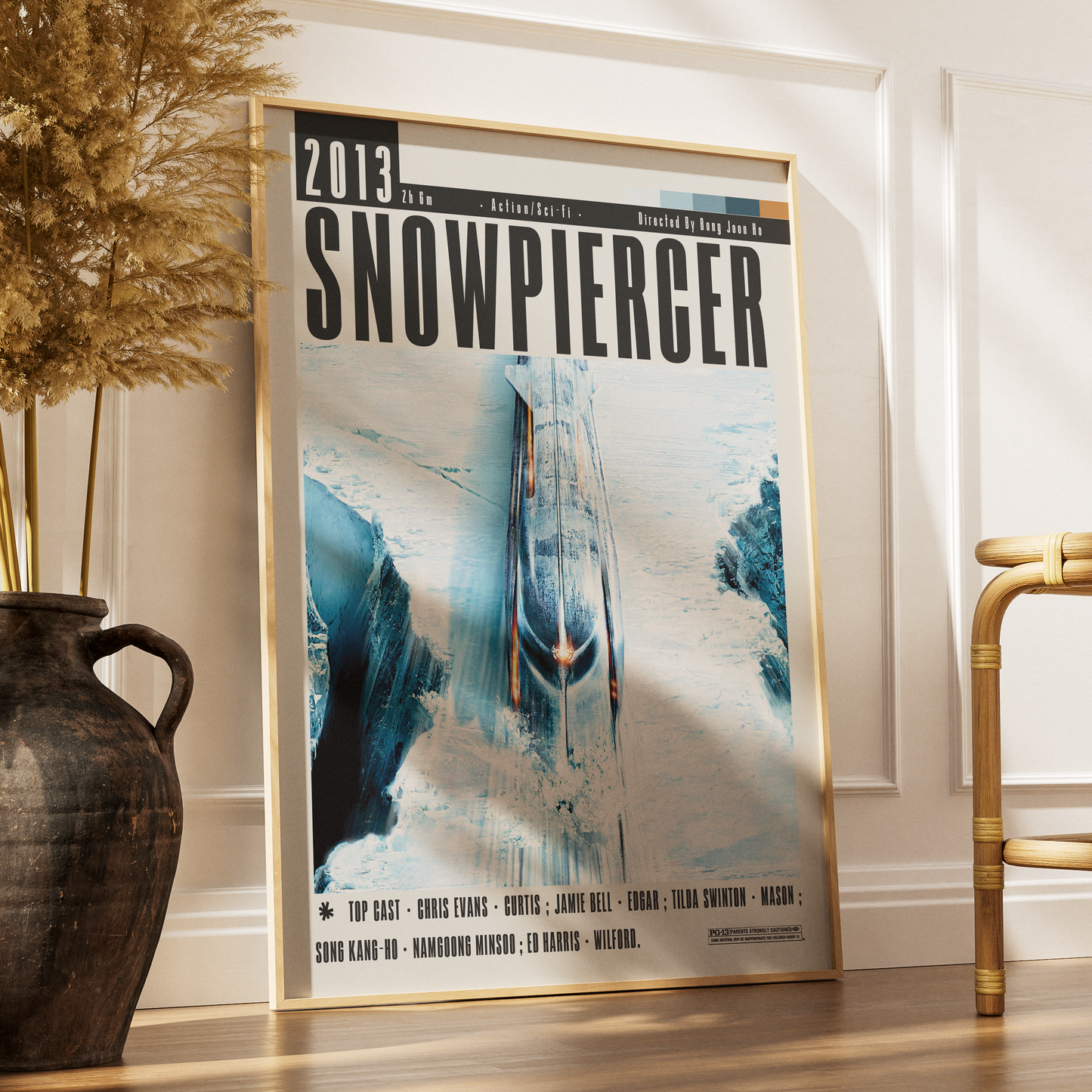 A framed Snowpierger movie poster leaning against a white wall on a wooden floor, styled in a room with a dark vase of dried grass on the left and a bamboo chair on the right.