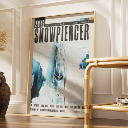 A framed Snowpiercer movie poster leaning against a white paneled wall in a styled interior setting with a wooden floor and a bamboo side table.