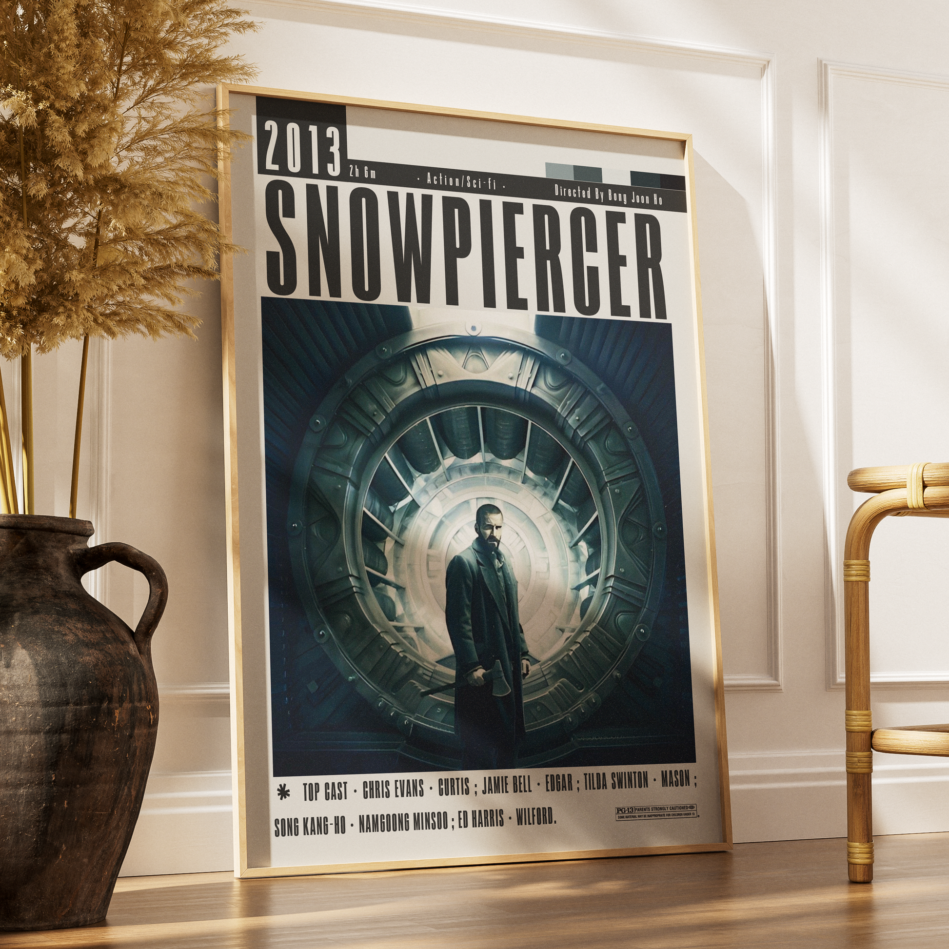 A framed Snowpierger movie poster leaning against a white wall on a wooden floor, styled in a home interior next to a dark ceramic vase with dried grass and part of a bamboo chair.