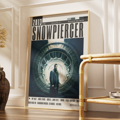 A framed Snowpiercer movie poster leaning against a white paneled wall on a wooden floor, with a woven chair and a decorative vase nearby.