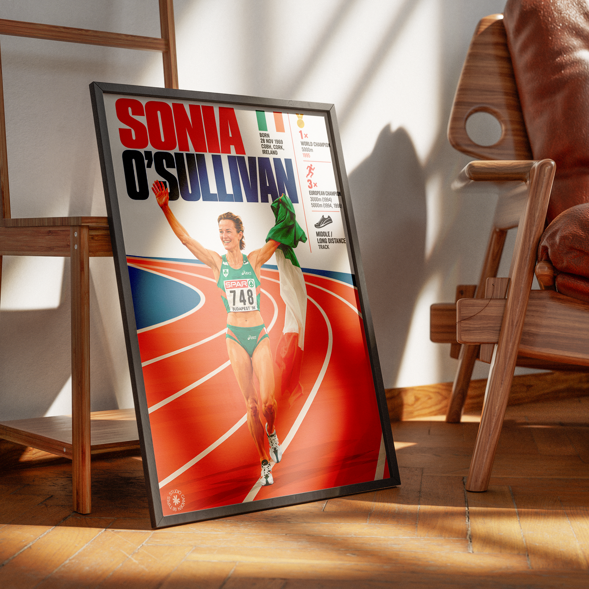 A framed poster of Sonia O'Sullivan leaning against a wall on a wooden floor, styled in a modern room with wooden chairs and warm sunlight casting shadows.