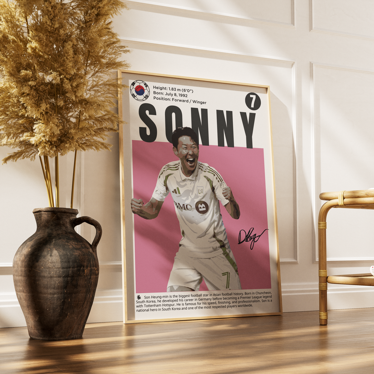 A framed sports poster of South Korean footballer Sonny Zhang leaning against a white paneled wall on a wooden floor, styled with a dark ceramic vase of dried grass on the left and a gold chair on the right.