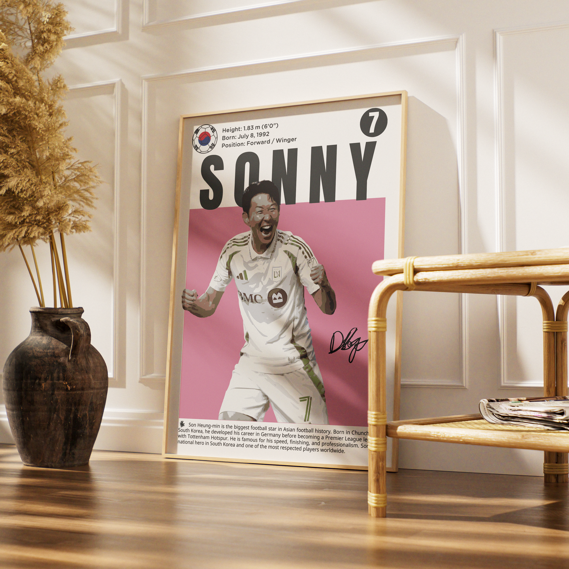 A framed sports poster of footballer Sonny Wagner leaning against a white paneled wall on a wooden floor, styled with a dark vase of dried grass and a bamboo side table.
