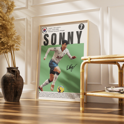 A framed poster featuring a vintage-style football player with the name SONNY and a South Korean flag emblem, leaning against a white paneled wall beside a rattan side table and a dark vase with dried grass.