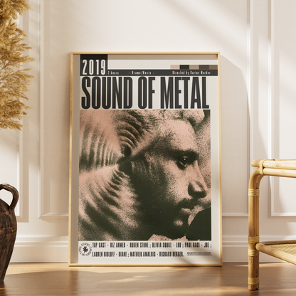 A framed music poster for the 2019 metal band Sound of Metal, leaning against a white paneled wall on a wooden floor, with a woven chair and dried plant visible in the room.
