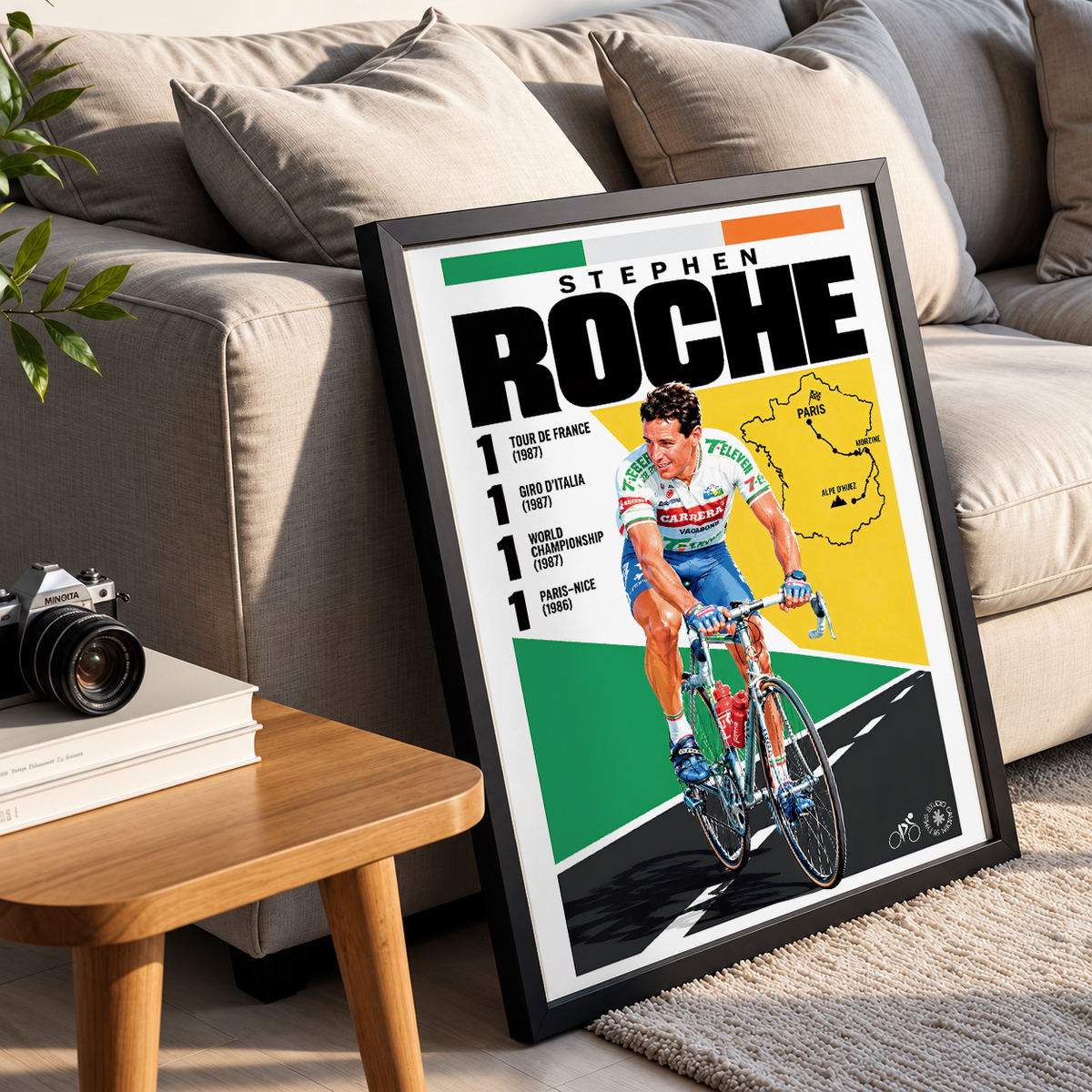 A framed cycling poster of Stephen Roche leaning against a beige sofa in a living room setting, with a small wooden side table and books in the foreground.