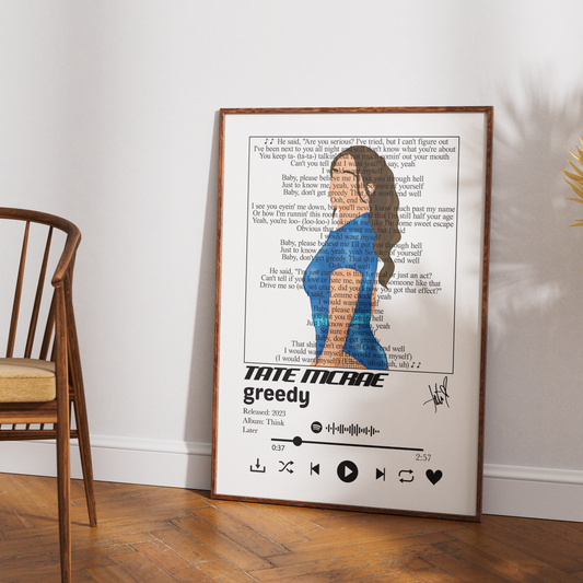 A framed music-themed art print of Tate McRae's song 'greedy' leaning against a white wall on a wooden floor, next to a wooden chair.