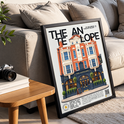 A framed art print leaning against a beige sofa in a living room setting. The print features a stylized illustration of The Astorhouse pub with the text 'THE ANTELOPE' and 'The Art of London 2024'.