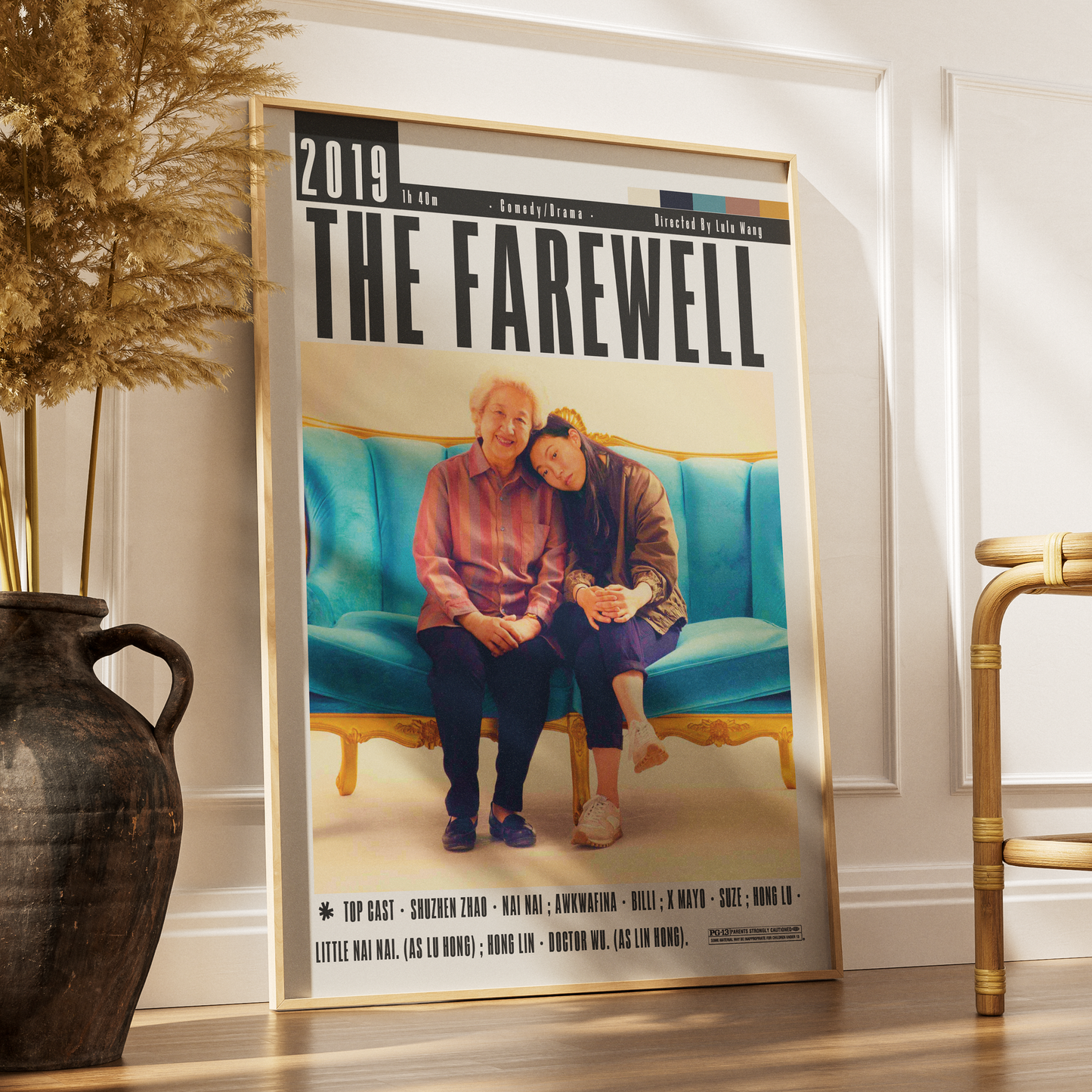 A framed vertical poster leaning against a white wall on a wood floor, next to a dark vase with dried grass and part of a gold chair. The poster reads '2019 The Farewell' and shows two people sitting on a turquoise sofa.