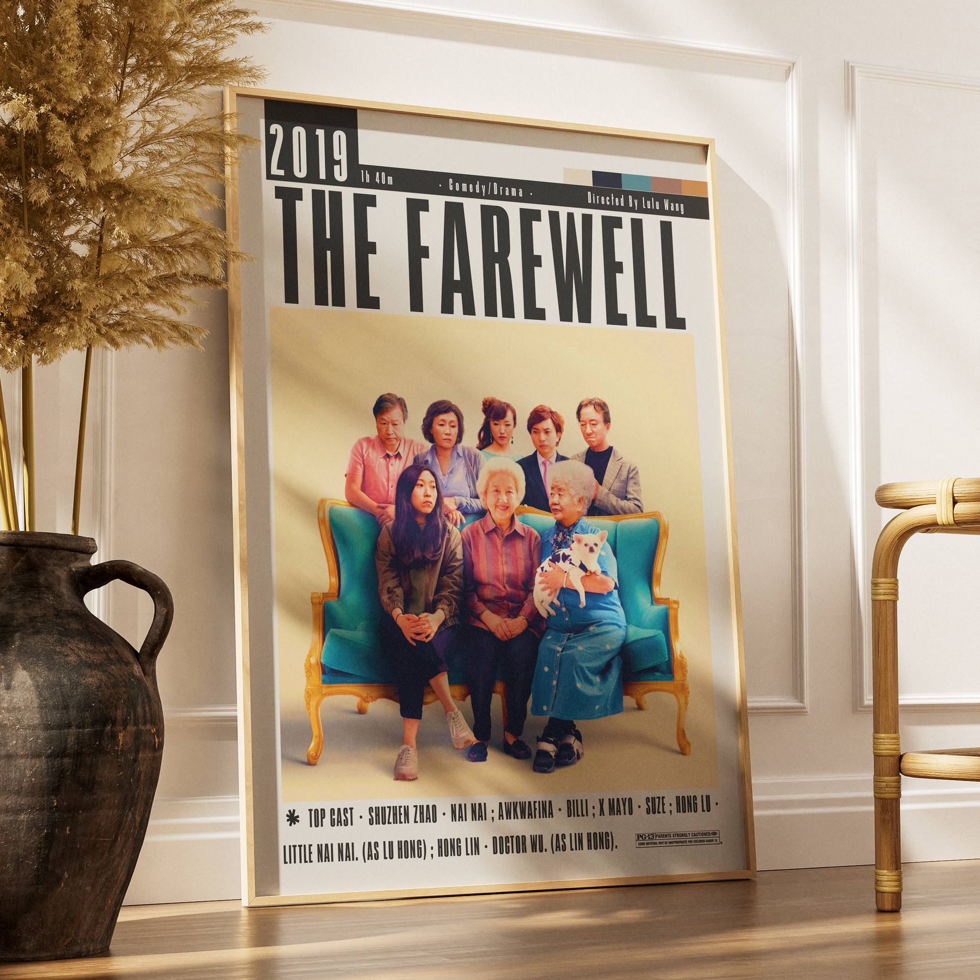 A framed movie poster for 'The Farewell' leaning against a white wall on a wooden floor, next to a dark vase with dried grass and part of a gold chair.