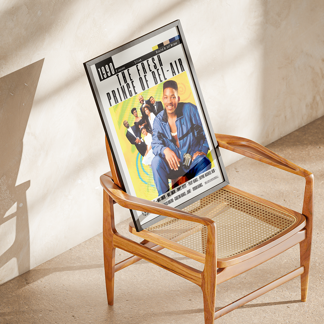 A framed 'The Fresh Prince of Bel-Air' movie poster leaning against a mid-century modern wooden chair with a woven cane seat, set in a sunlit room with textured plaster walls.