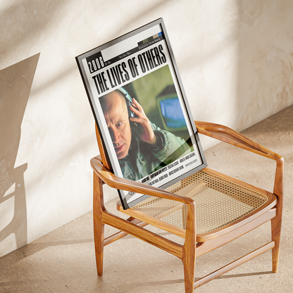 A framed art print titled 'The Lives of Others' displayed on a mid-century modern wooden armchair with a woven cane seat, set against a textured beige plaster wall with warm sunlight and shadows.