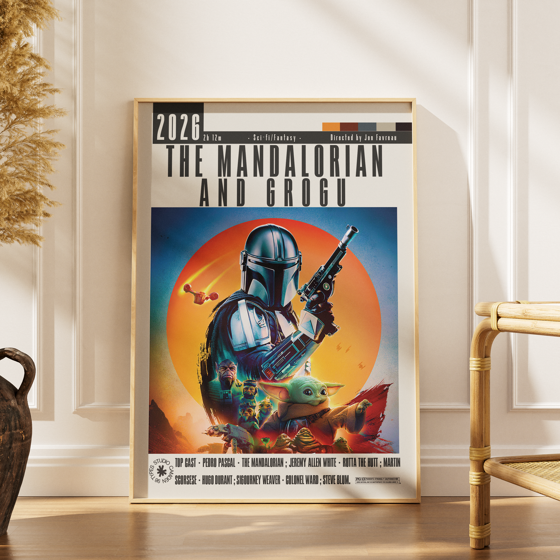 A framed vertical poster of 'The Mandalorian and Grogu' styled on a wooden floor against a cream paneled wall, with a dried plant on the left and a rattan chair on the right.
