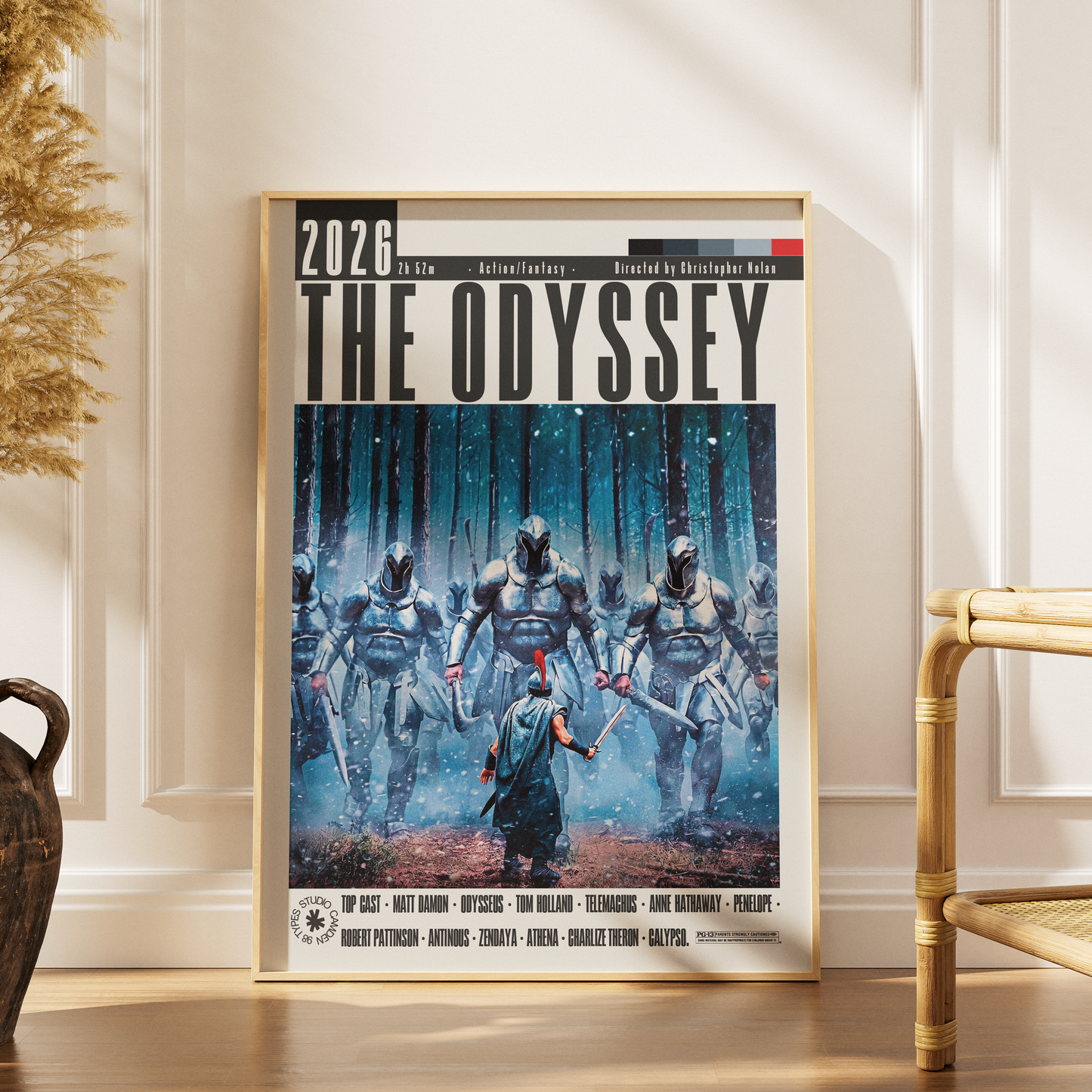 A framed vertical poster titled "THE ODYSSEY" leaning against a cream wall on a wood floor, with a bamboo chair and dried plant visible in the room. The poster artwork shows armored figures in a blue-toned forest scene with a central figure in blue and red at the bottom.