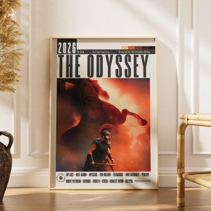 A framed movie poster for 'The Odyssey' (2025) leaning against a white paneled wall on a wooden floor, next to a woven chair and dried grass.
