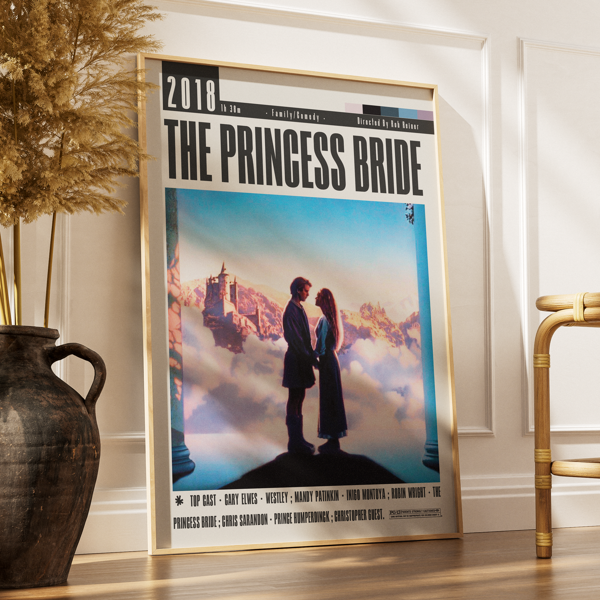 A framed vertical poster of the movie 'The Princess Bride' leaning against a white wall on a wooden floor, styled in a room with a dark vase of dried grass on the left and a gold chair on the right.