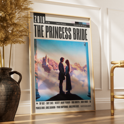 A framed vertical poster of the movie 'The Princess Bride' leaning against a white wall on a wooden floor, styled in a room with a dark vase of dried grass on the left and a gold chair on the right.