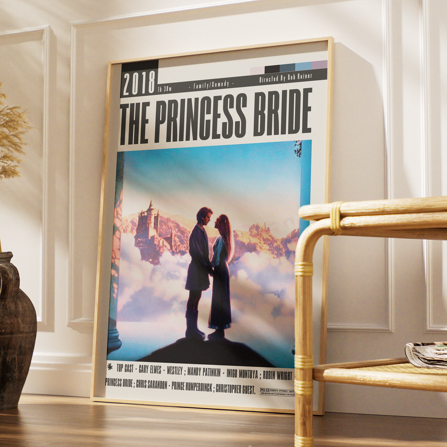 A framed movie poster for 'The Princess Bride' leaning against a white paneled wall in a styled interior setting with a wooden floor and a rattan chair partially visible.