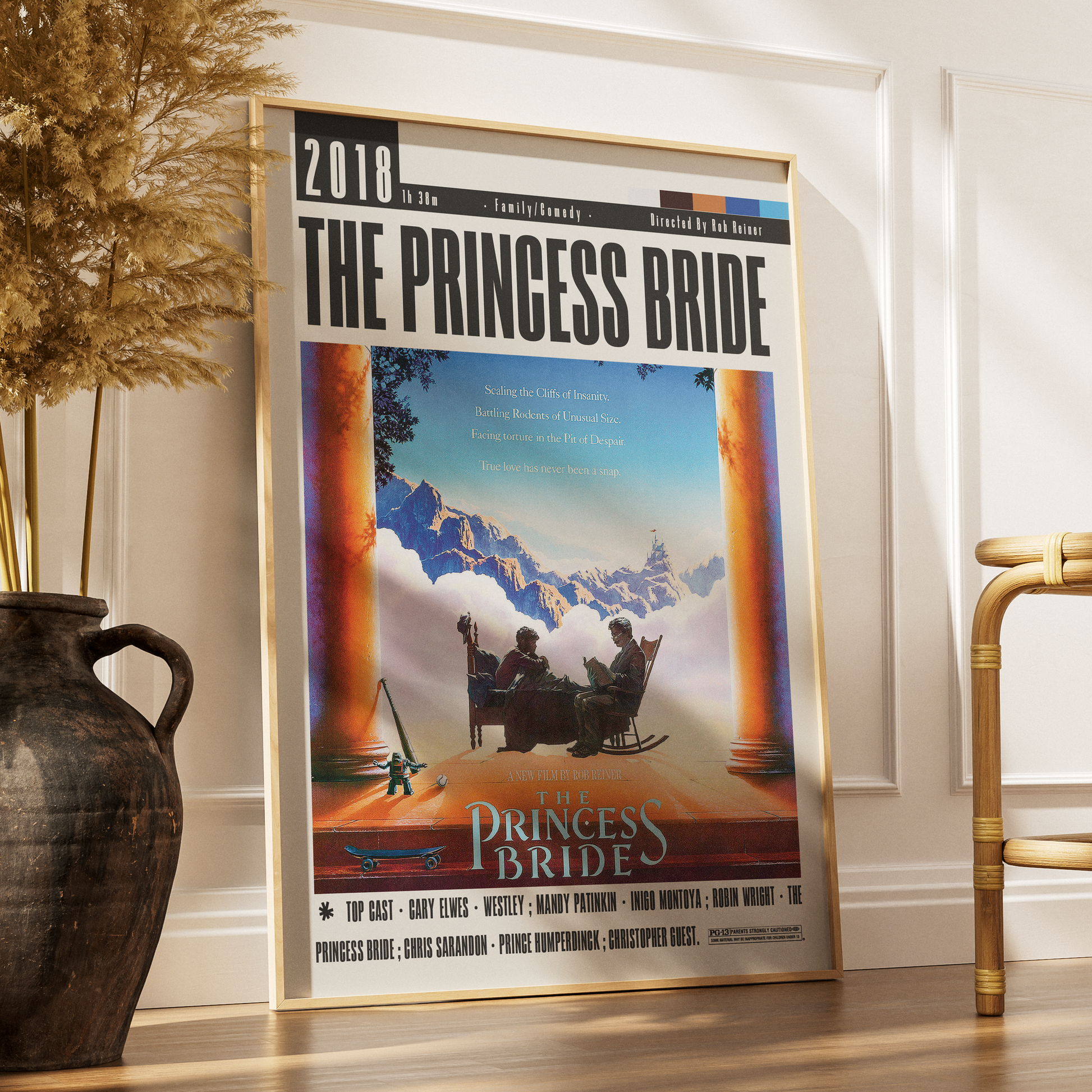 A framed vertical art print of Disney's 'The Princess Bride' leaning against a white wall on a wooden floor, styled in a room with a dark ceramic vase and dried grass on the left and a gold chair on the right.