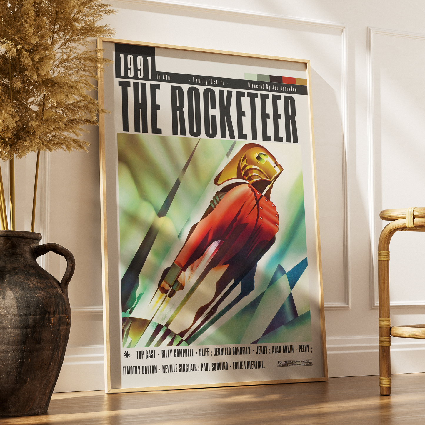 A framed vintage-style poster titled "THE ROCKETEER" leaning against a white wall on a wooden floor, with a dark vase of dried grass on the left and part of a chair on the right.
