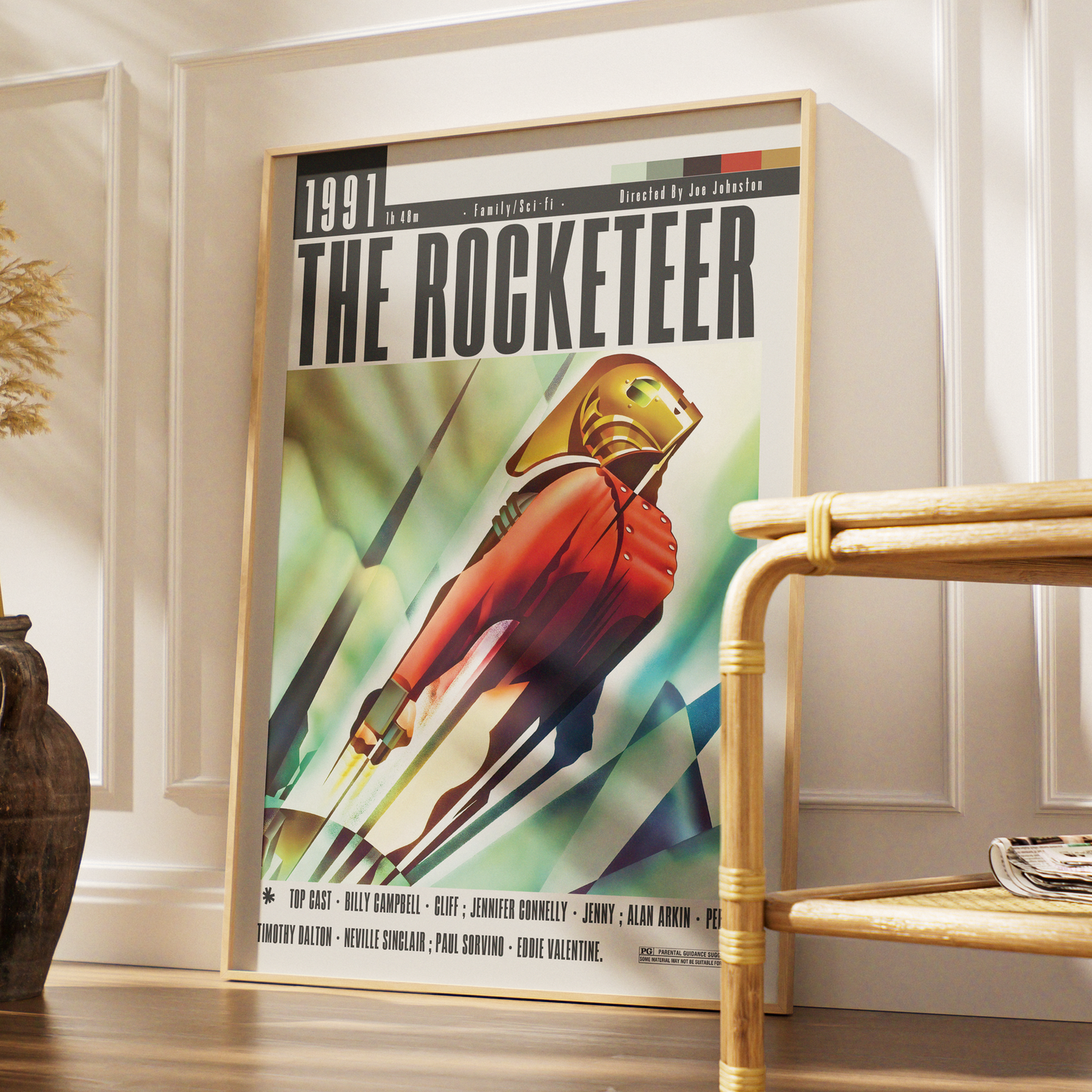 A framed vintage-style poster titled "THE ROCKETEER" leaning against a white paneled wall in a styled interior. The poster shows a red-suited astronaut or space figure with a gold helmet against a green and blue abstract background.