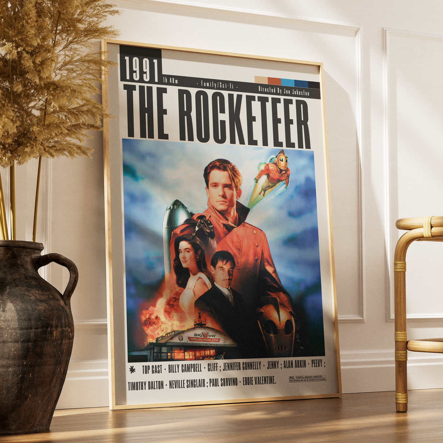 A framed movie poster for 'The Rocketeer' leaning against a white wall on a wooden floor, styled in a room with a dark ceramic vase and dried grass on the left and a gold chair on the right.