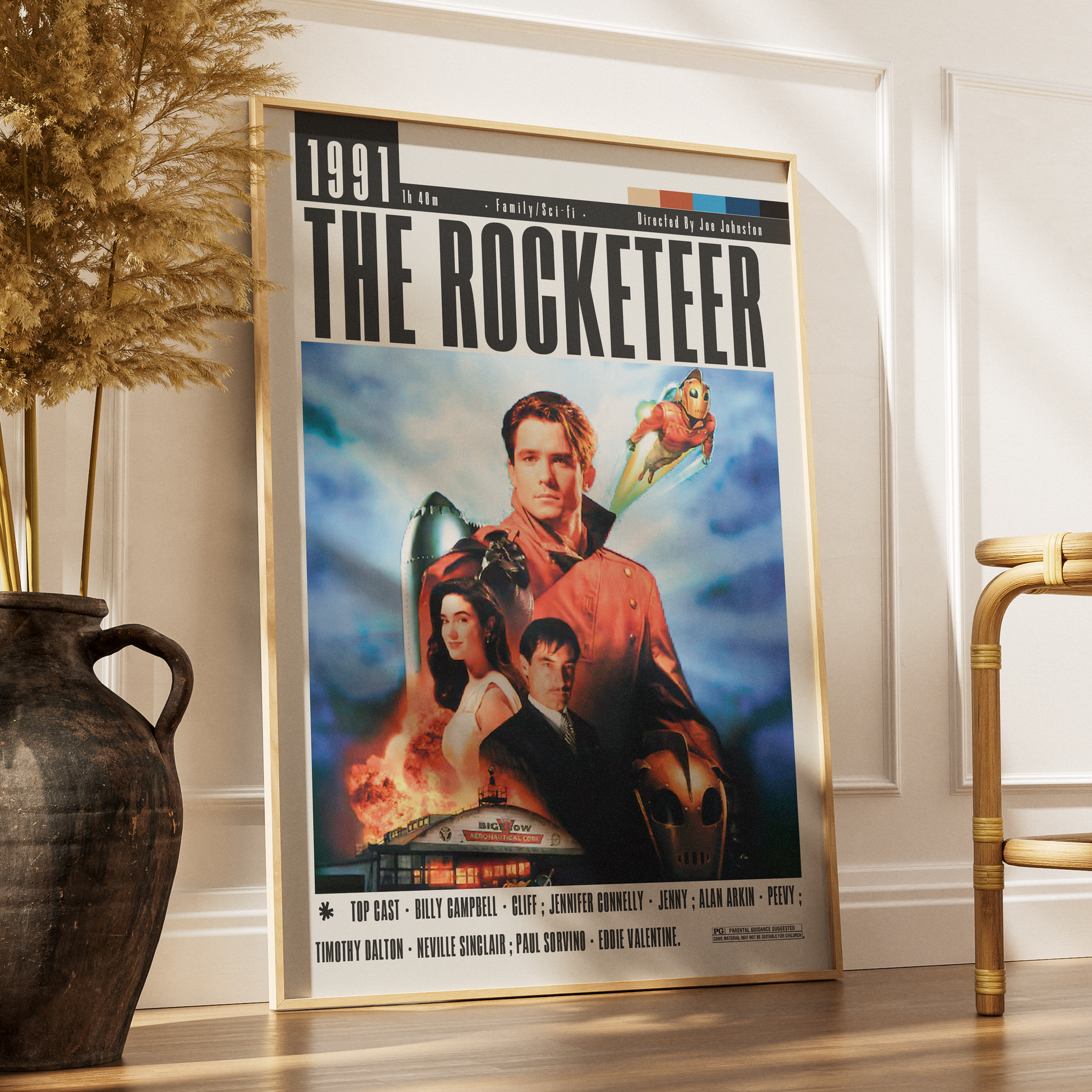 A framed movie poster for 'The Rocketeer' leaning against a white wall on a wooden floor, styled in a room with a dark ceramic vase and dried grass on the left and a gold chair on the right.