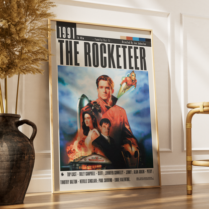 A framed movie poster for 'The Rocketeer' leaning against a white wall on a wooden floor, styled in a room with a dark ceramic vase and dried grass on the left and a gold chair on the right.