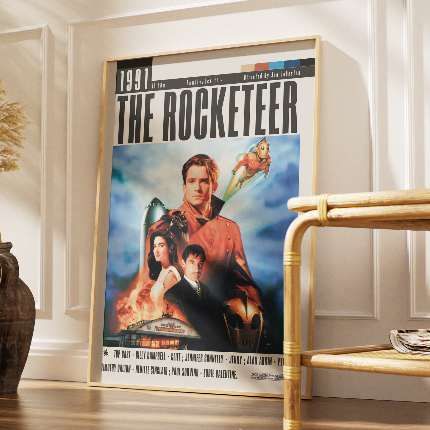A framed movie poster for 'The Rocketeer' leaning against a white paneled wall in a styled interior setting with a wooden floor and a bamboo side table.