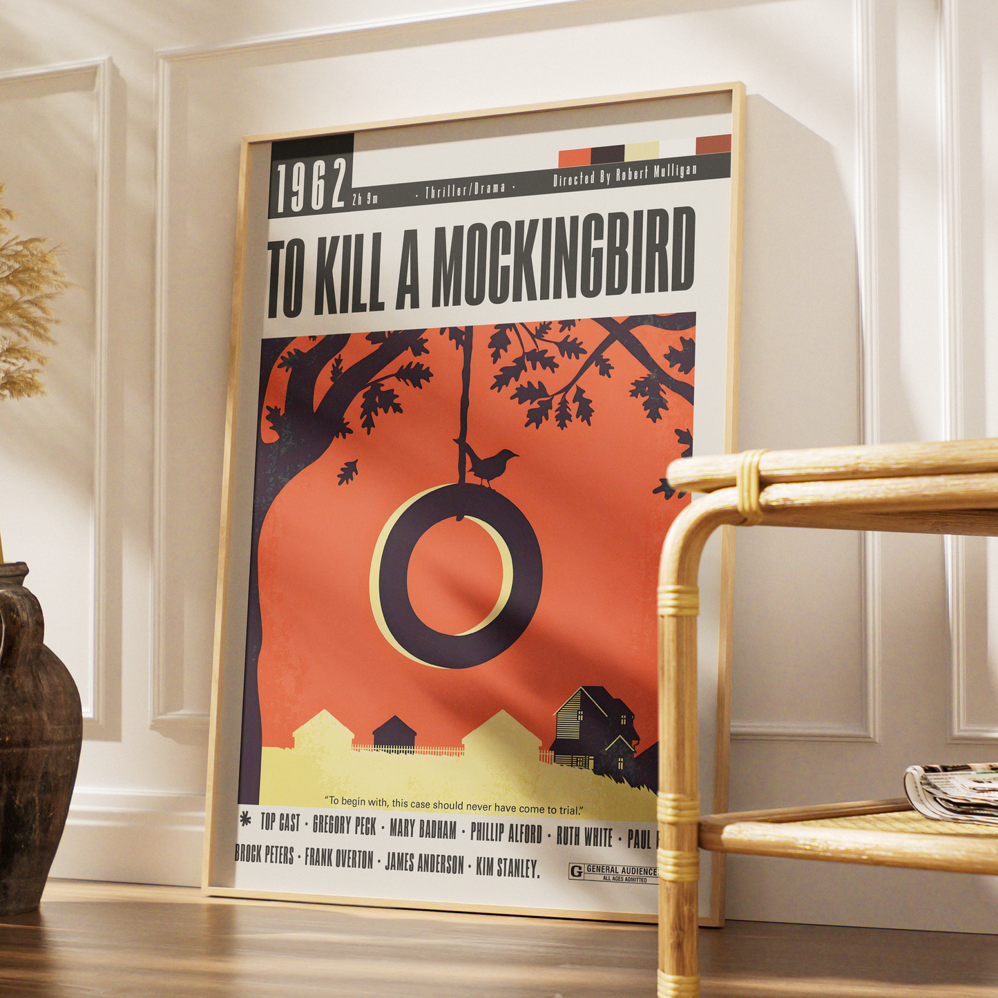 A framed movie poster for 'To Kill a Mockingbird' leaning against a white paneled wall in a styled interior setting with a bamboo side table and a dark vase.
