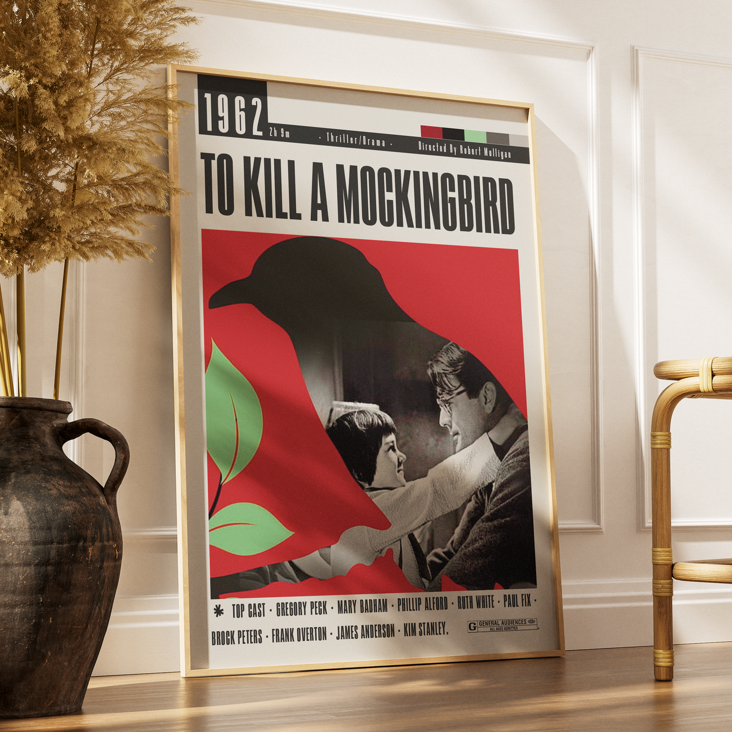 A framed vintage-style movie poster for 'To Kill a Mockingbird' leaning against a white wall on a wooden floor, with a dark vase and dried grass on the left and part of a chair on the right.